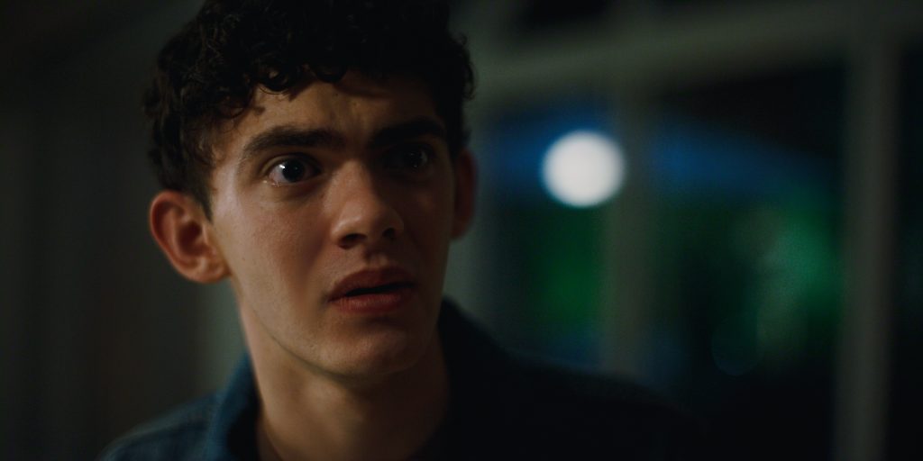 "Young man with curly hair looking surprised or anxious indoors at night, with blurred windows and bright light visible in the background."