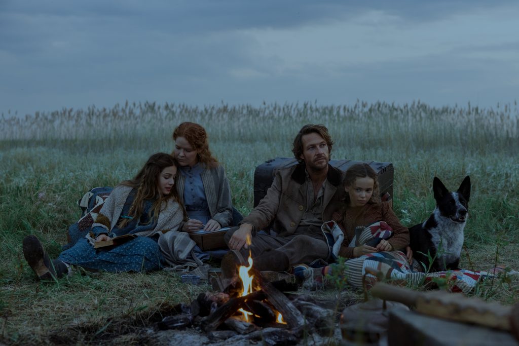"A family of four and a dog sit together by a campfire in a grassy field under a cloudy sky, surrounded by camping gear, suggesting a rustic outdoor setting and a calm, intimate atmosphere."