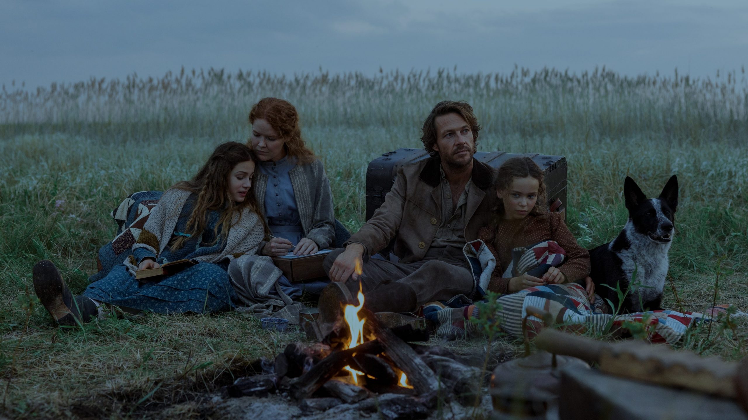 "A family of four and a dog sit together by a campfire in a grassy field under a cloudy sky, surrounded by camping gear, suggesting a rustic outdoor setting and a calm, intimate atmosphere."