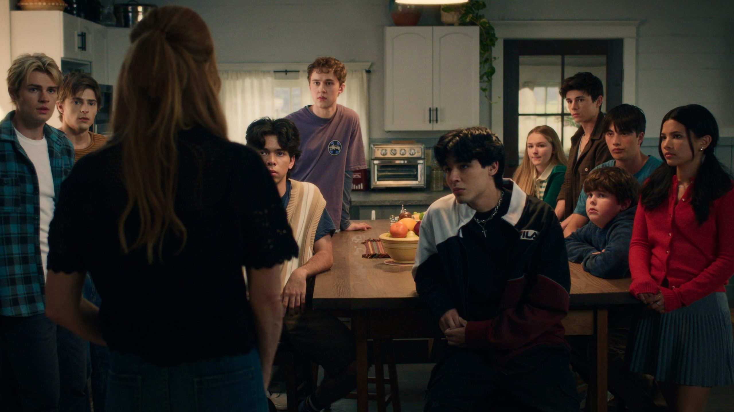 "A woman stands facing a group of teenagers gathered around a kitchen table in a cozy, warmly lit home, all focused on her as if listening intently to an important conversation."