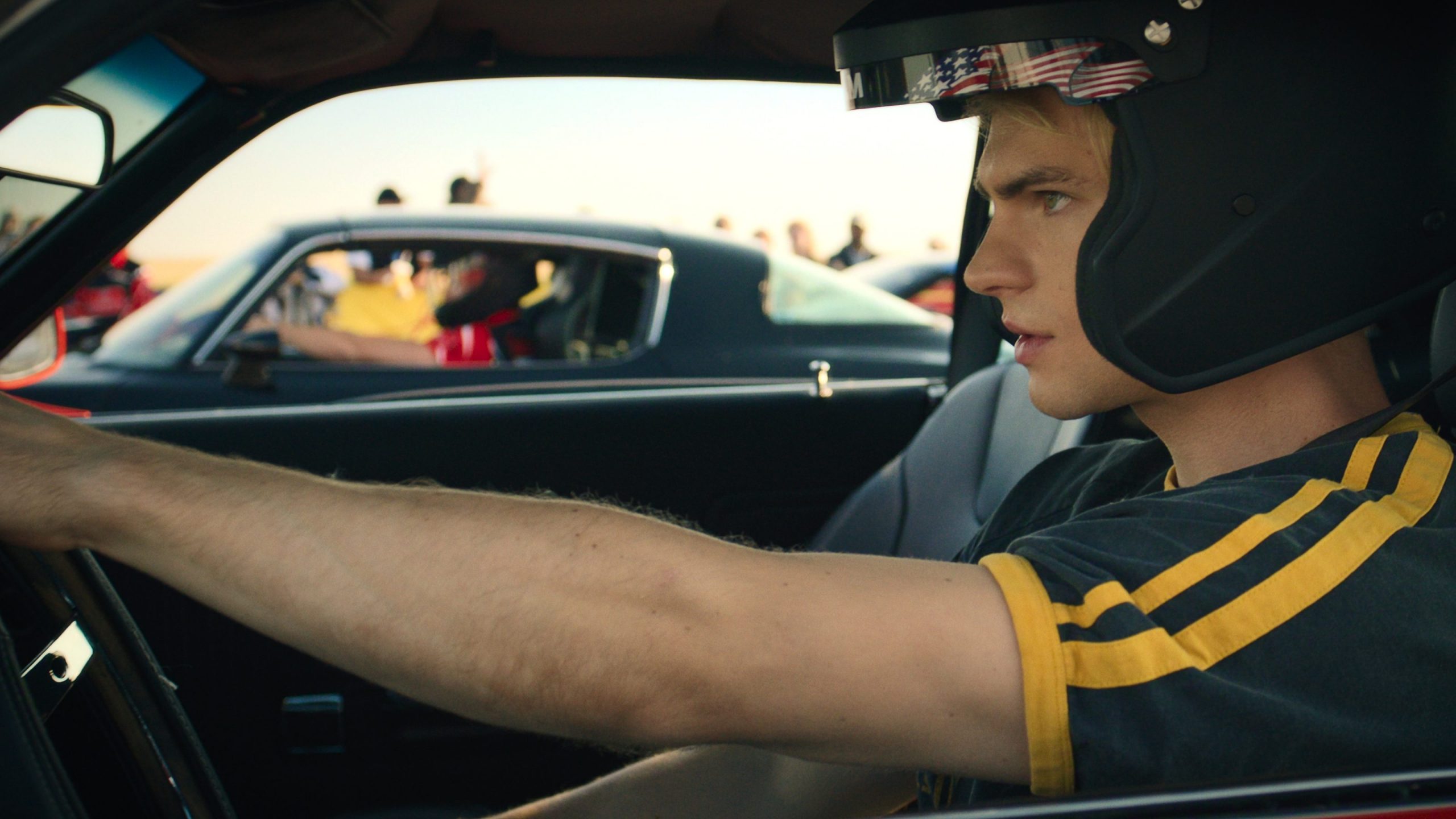 "Young man wearing a racing helmet sitting in a car, gripping the steering wheel, preparing for a race. Other cars and people are visible in the background, suggesting a motorsport or racing event under daylight."