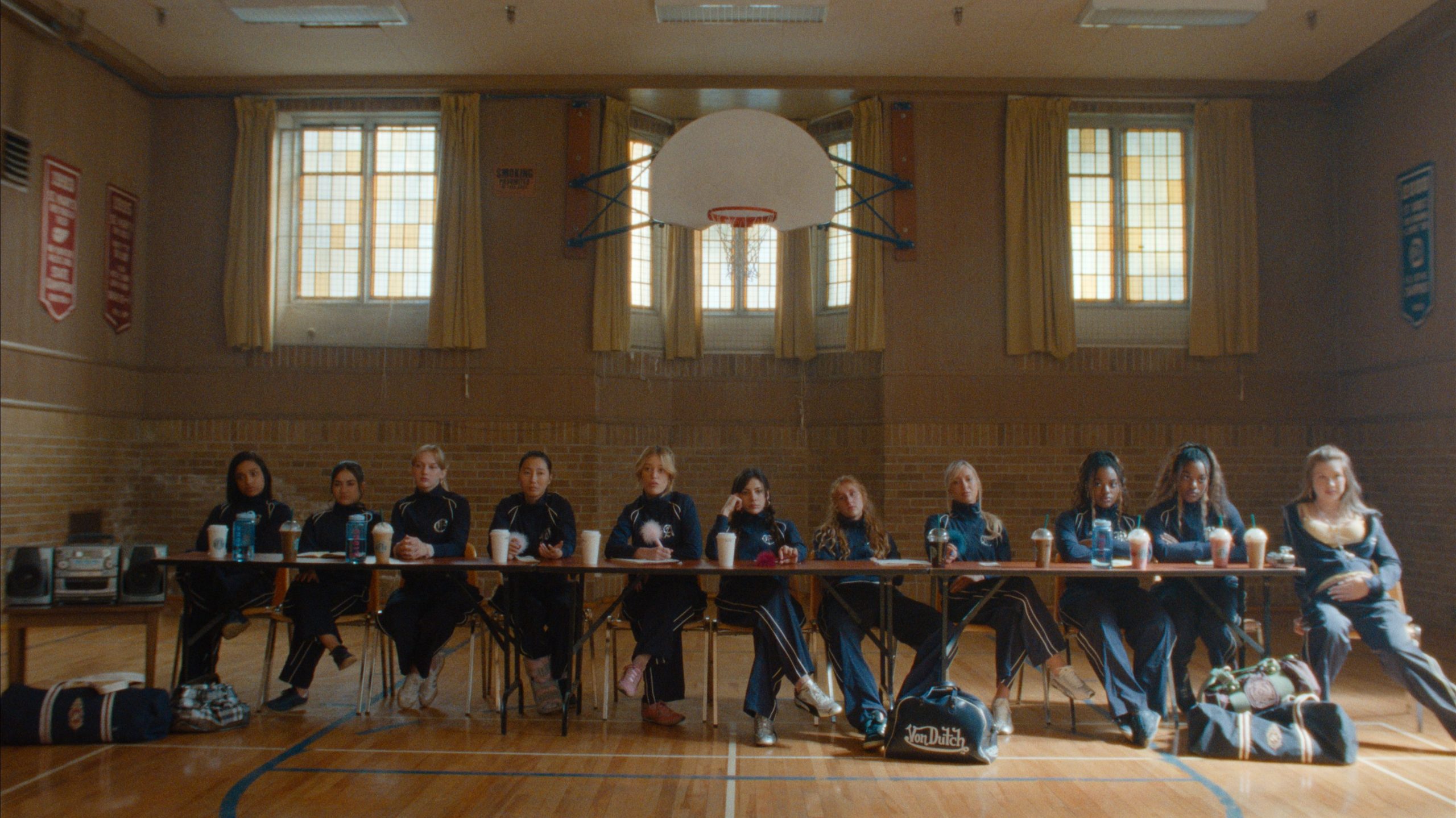 "A group of girls in athletic uniforms sit behind a long table in a gymnasium, with sports bags on the floor, under a basketball hoop and surrounded by tall windows and wall banners."