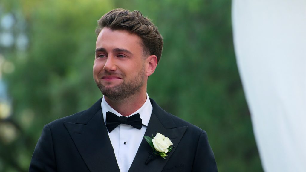 "A man in a black tuxedo with a white rose boutonniere stands outside, smiling with greenery in the background, likely at a formal event such as a wedding."