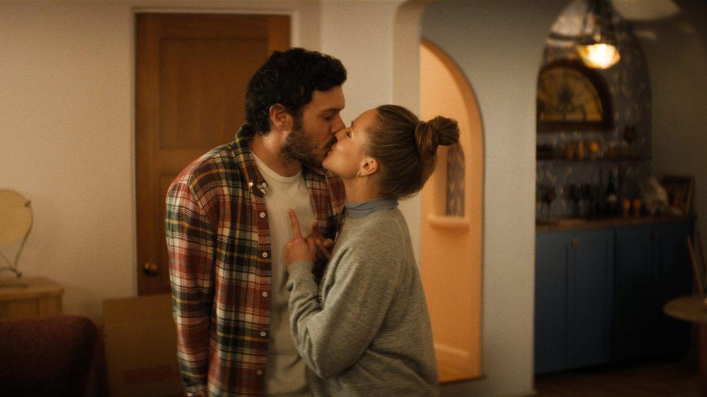 "A man and woman stand close together in a cozy, warmly lit living room, appearing to argue or have an intense conversation near a wooden door and a kitchen alcove with blue cabinets in the background."
