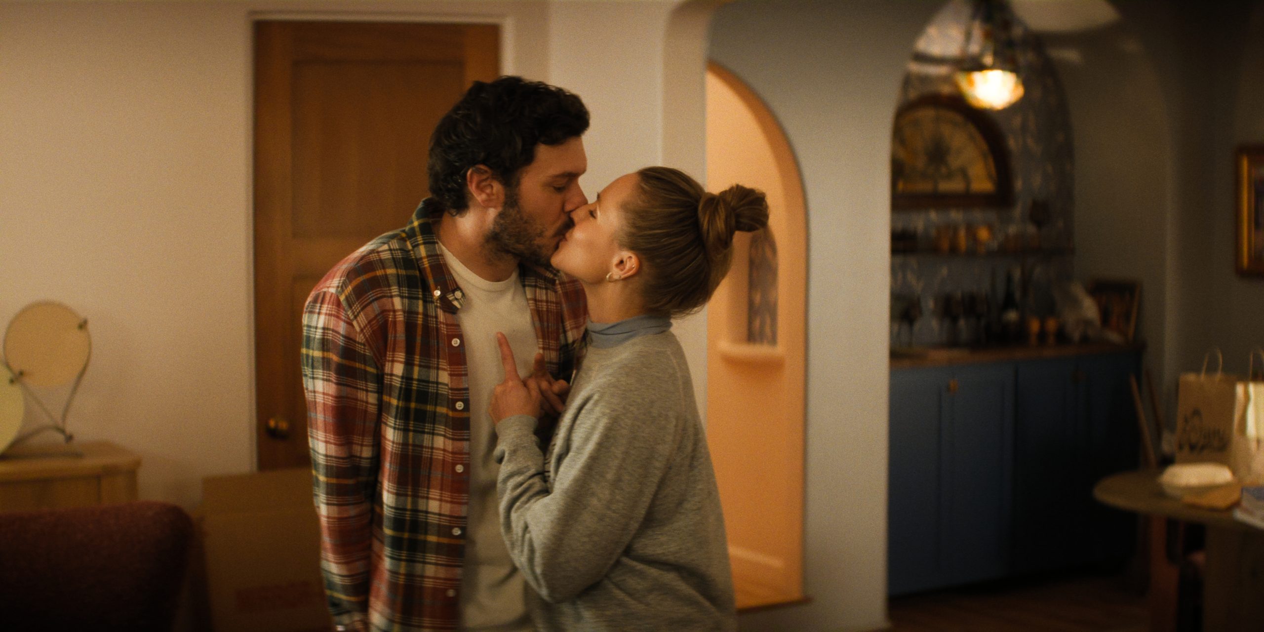 "A man and woman stand close together in a cozy, warmly lit living room, appearing to argue or have an intense conversation near a wooden door and a kitchen alcove with blue cabinets in the background."