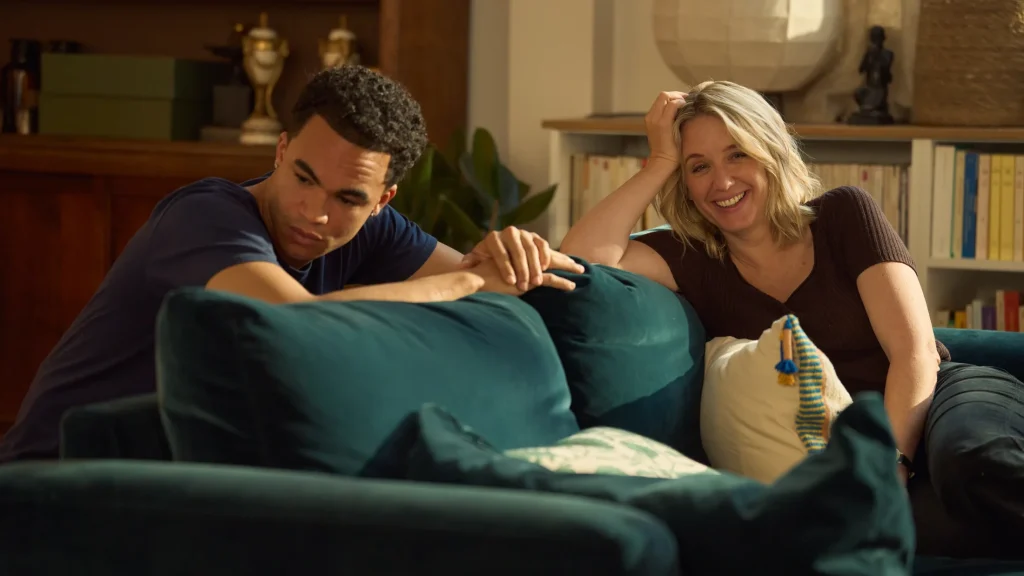 "Two people sitting on a teal sofa in a cozy living room. The man looks thoughtful while the woman smiles warmly. Shelves with books, plants, and decor are in the background, creating a homey atmosphere with soft natural light."